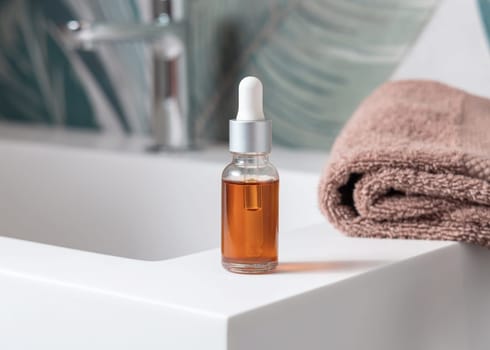 Glass dropper bottle with pipette filled with orange liquid near brown towels and faucet on basin in green tropical bathroom close up. Natural cosmetics - serum or essence. Spa concept