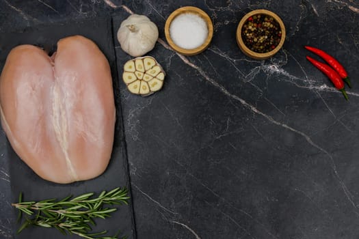 Chicken breast fillet lies on a black board on a dark background with a sprig of rosemary next to it, garlic, red pepper and spices and salt on top in wooden bowls with space for text. The concept of proper and healthy nutrition, dietary dishes and recipes. High quality photo
