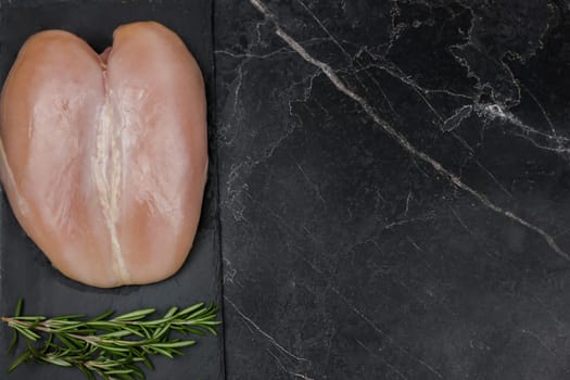 Chicken breast fillet lying on a black board on a dark background with a sprig of rosemary on a dark background with space for text. The concept of proper and healthy nutrition, dietary dishes and recipes. High quality photo