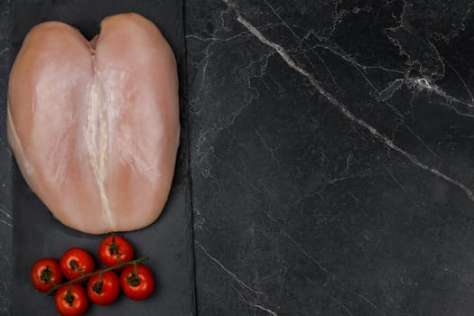 The chicken breast fillet lies on a black board on a dark background with a sprig of tomatoes next to the text space. The concept of proper and healthy nutrition, dietary dishes and recipes. High quality photo