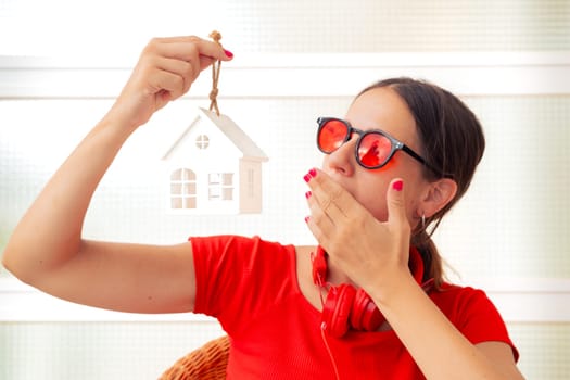 A young woman wearing red sunglasses and headphones holds a small house model while covering her mouth in surprise, displaying an expressive and curious gesture.