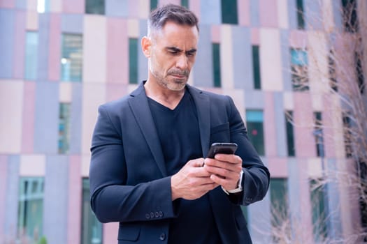 Smiling middle-aged businessman in a blue shirt holding and using a smartphone outdoors in a modern urban setting.