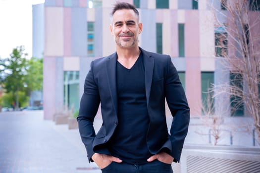 Smiling businessman in Mature professional man wearing a black suit, standing confidently outdoors with a serious expression. Modern city architecture emphasizes corporate strength and authority.