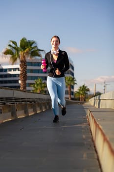 A smiling fitness woman runs with headphones and water bottle outdoors. Workout concept and healthy lifestyle.
