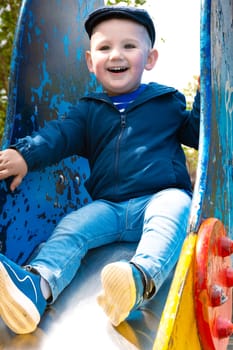 Sweet little Caucasian boy in cap having fun smiling widely sliding down the slide.