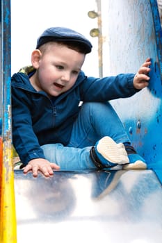 Sweet little Caucasian boy in cap having fun smiling widely sliding down the slide.