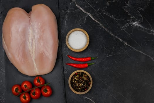 Chicken breast fillet lies on a black board on a dark background with a sprig of tomatoes next to it, spices and salt in wooden bowls on the side, red hot pepper on a dark background with space for text. The concept of proper and healthy nutrition, dietary dishes and recipes. High quality photo