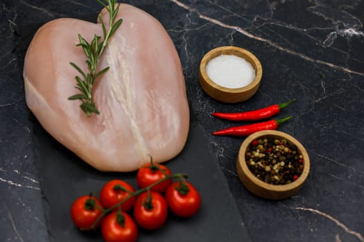 Close-up of chicken breast fillet lying on a black board on a dark background with a sprig of tomatoes next to it, spices and salt in wooden bowls on the side, red hot pepper on a dark background. The concept of proper and healthy nutrition, dietary dishes and recipes. High quality photo