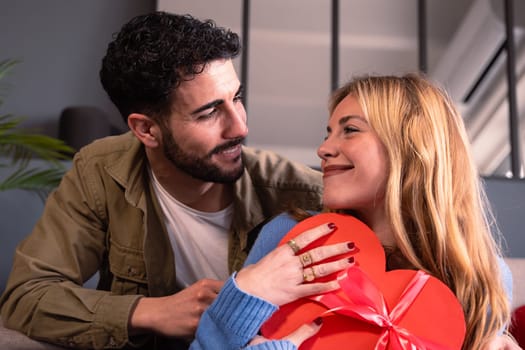 Beautiful young couple at home enjoying time together while celebrating Valentine's Day with a heart-shaped gift box in hand. Looking at each other with love.