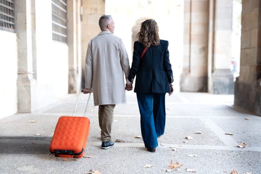 Smiling middle-aged Caucasian tourist couple walking hand-in-hand carefree with their luggage looking at each other. Happy husband and wife enjoying their outdoor vacation on a sunny winter day