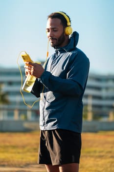 Athletic man with headphones tired from exercising takes a break to drink water while using a cell phone app outdoors.