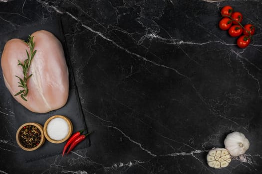 Chicken breast fillet with spices and salt in wooden bowls and red hot pepper lies on a black board on the other side with a sprig of tomato and garlic on a dark background with space for text in the center. The concept of proper and healthy nutrition, dietary dishes and recipes. High quality photo