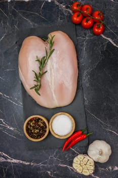 Close-up in the center of chicken breast fillet with spices and salt in wooden bowls and red hot pepper lying on a black board, next to a sprig of tomato and garlic on a dark background. The concept of proper and healthy nutrition, dietary dishes and recipes. High quality photo