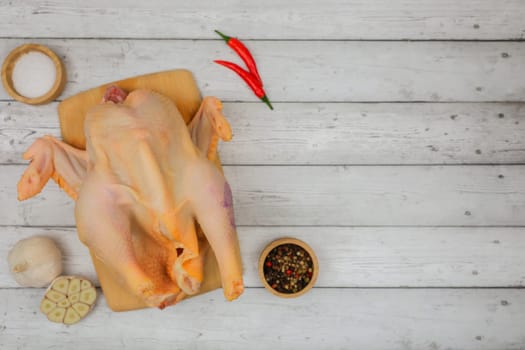 A raw processed chicken carcass lies on a white wooden background on a wooden board with spices and salt in wooden bowls, garlic and red pepper with space for text. The concept of cooking, wholesome and healthy food, recipes. High quality photo