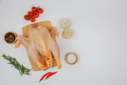 The raw processed chicken carcass lies on a wooden board with spices in wooden bowls, garlic and red pepper, tomatoes and rosemary with space for text on a white background. The concept of cooking, wholesome and healthy food, recipes. High quality photo