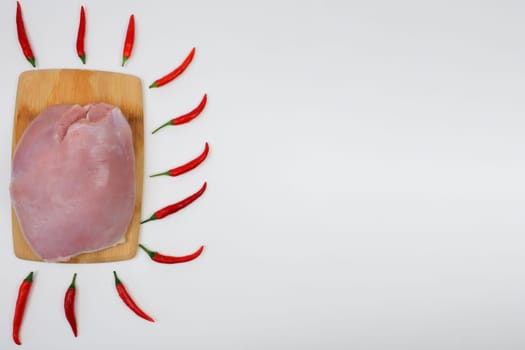 The raw turkey fillet lies on a wooden cutting board, surrounded by red hot chili peppers on a white background with space for text. The concept of dietary dishes, spicy food, healthy and natural products. High quality photo
