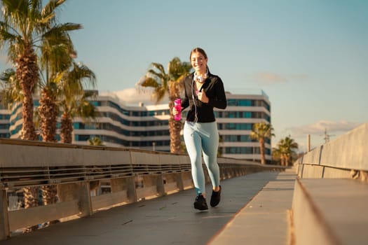 A happy fitness woman runs with headphones and water bottle looking at camera. Workout concept and healthy lifestyle.
