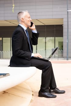 A middle-aged adult man in a suit has a business call while looking at his laptop.