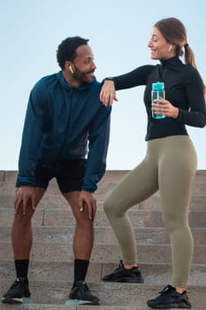 Smiling sporty young couple with sports bottle, taking a break on the stairs while exercising outdoors.