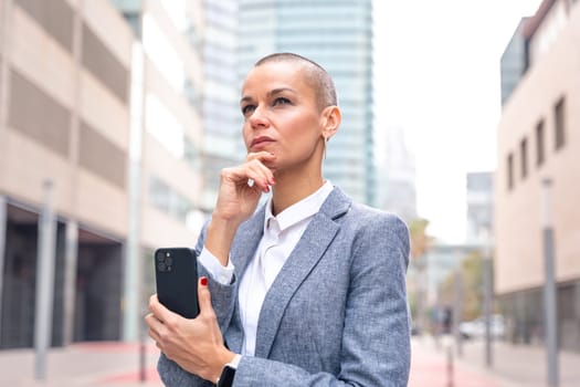 Thoughtful businesswoman with results of businesswoman walking down the street outside office building, a mature boss holds a phone in her hands, writes messages and reads news online, using an app.