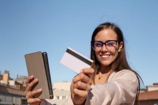 Happy young woman with glasses holding in each hand a credit card and digital tablet. Looking into the camera. Concept: payment in an online store