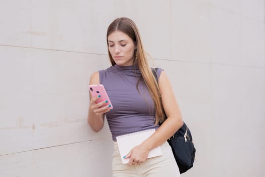 Front view of a laughing Caucasian teenage girl using a social media application with her mobile phone, leaning on the wall. University life concept
