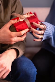 Elegant man holding gift with ribbon and giving it to his girlfriend on the sofa at home. Celebrating Valentine's Day