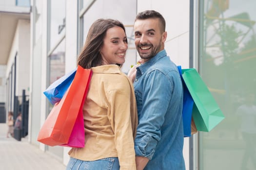 Young happy couple walking with shopping bags and turning back smiling to look and wave at the camera on an urban city street. Consumption concept.