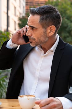 Middle aged man sitting drinking coffee happily having a work conversation with cell phone.