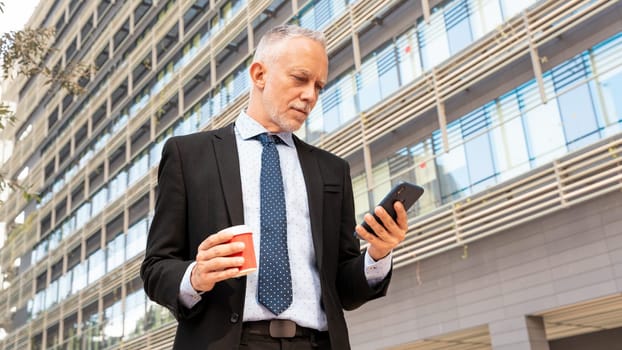 Satisfied with the results, the businessman walks down the street outside the office building, a mature boss holds a phone and coffee in his hands, writes messages and reads news online, using an app.