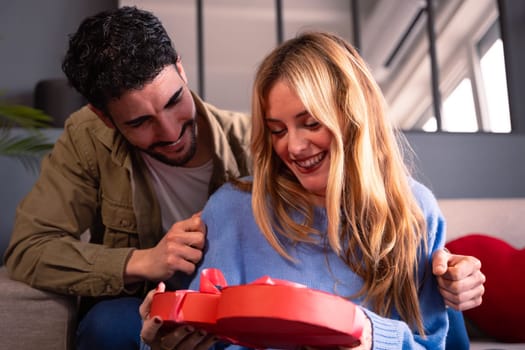Lovely young couple at home enjoying time together while celebrating Valentine's Day with a heart-shaped gift box in hand.