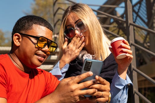 Happy diverse friends in the park sitting together on a bench surprised looking at social media on smartphones talking and smiling.