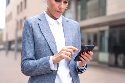 Satisfied with the results, the businesswoman walking on the street outside the office building, a mature boss holds a phone in her hands, writes messages and reads news online, using an app.