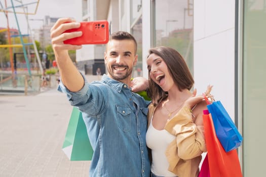 Couple With Shopping Bags Making Selfie On Cell Phone Having Fun Standing In Hypermarket. Spouses Enjoy Weekend In Modern Shopping Center. Black Friday sale, seasonal offer advertising concept