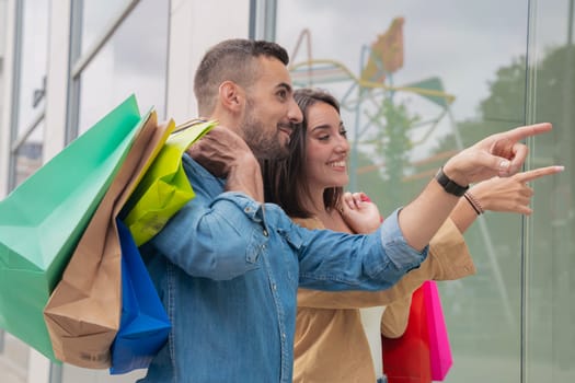 happy young couple carrying shopping bags shopping in mall pointing new clothes in shop window, showing good discount price offer.