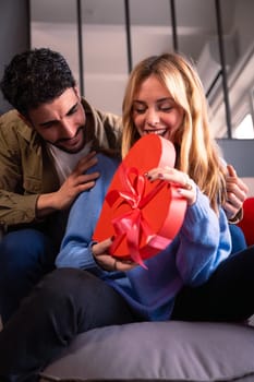 Beautiful young couple at home enjoying time together while celebrating Valentine's Day with a heart-shaped gift box in hand