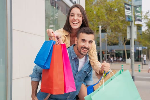 Happy caucasian man carrying his girlfriend on his back having fun together carrying shopping bags Concept of excitement, weekend and family.