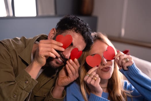 Smiling couple holding heart shapes over their eyes while sitting on the couch at home .Valentine's Day love.