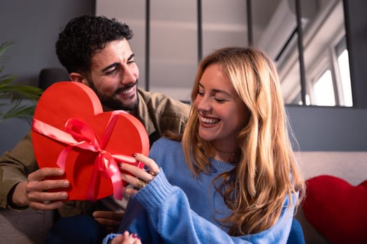 Beautiful young couple at home enjoying time together while celebrating Valentine's Day with a heart-shaped gift box in hand
