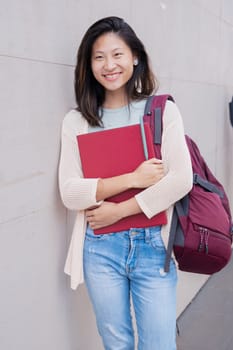 Front view of smiling Asian teenage girl with backpack leaning on the wall.College life concept
