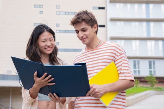 Happy university student friends talking and laughing while reviewing syllabus notes on campus. Diverse Asian and European youth