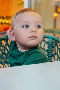 Close-up portrait of a very beautiful boy with blonde hair and big green eyes sitting on the terrace in a chair.