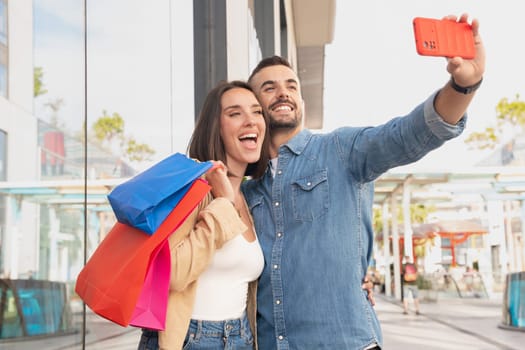 Couple With Shopping Bags Making Selfie On Cell Phone Having Fun Standing In Hypermarket. Spouses Enjoy Weekend In Modern Shopping Center. Black Friday sale, seasonal offer advertising concept