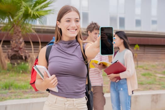 Young smiling student holding and showing an empty smartphone screen, behind her classmates talking and reviewing class notes.