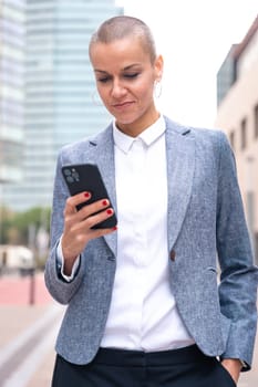 Satisfied with the results, the businesswoman walking on the street outside the office building, a mature boss holds a phone in her hands, writes messages and reads news online, using an app.