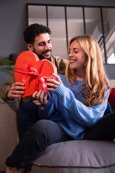 Beautiful young couple at home enjoying time together while celebrating Valentine's Day with a heart-shaped gift box in hand. Looking at each other with love.