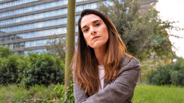 Caucasian business woman looking seriously at camera in a suit sitting in an outdoor workspace. Front view of a young lawyer with thoughtful expression watching.