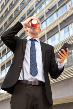Satisfied with the results, the businessman walks down the street outside the office building, a mature boss holds a phone and coffee in his hands, writes messages and reads news online, using an app.