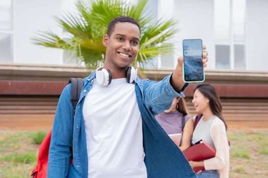 Young smiling black student holding and showing an empty smartphone screen, behind her classmates talking and reviewing class notes.
