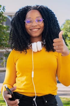 Close-up of happy curly haired young woman with glasses pointing thumb up. Facial expression of joy.